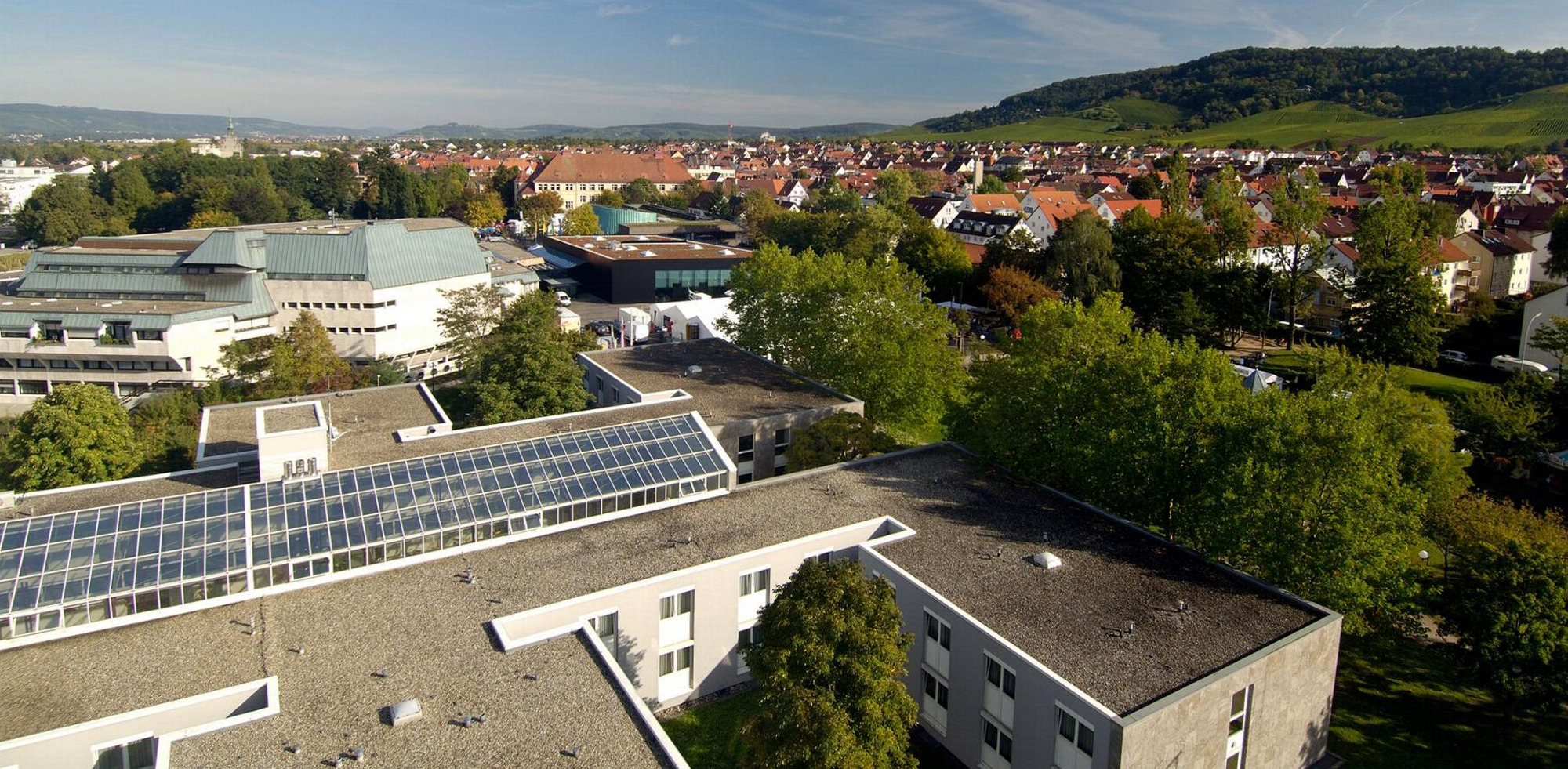 Panorama eines Wohngebiets mit Häusern, Bäumen, Hügeln und Glasdach im Vordergrund, teils bewölkter Himmel.