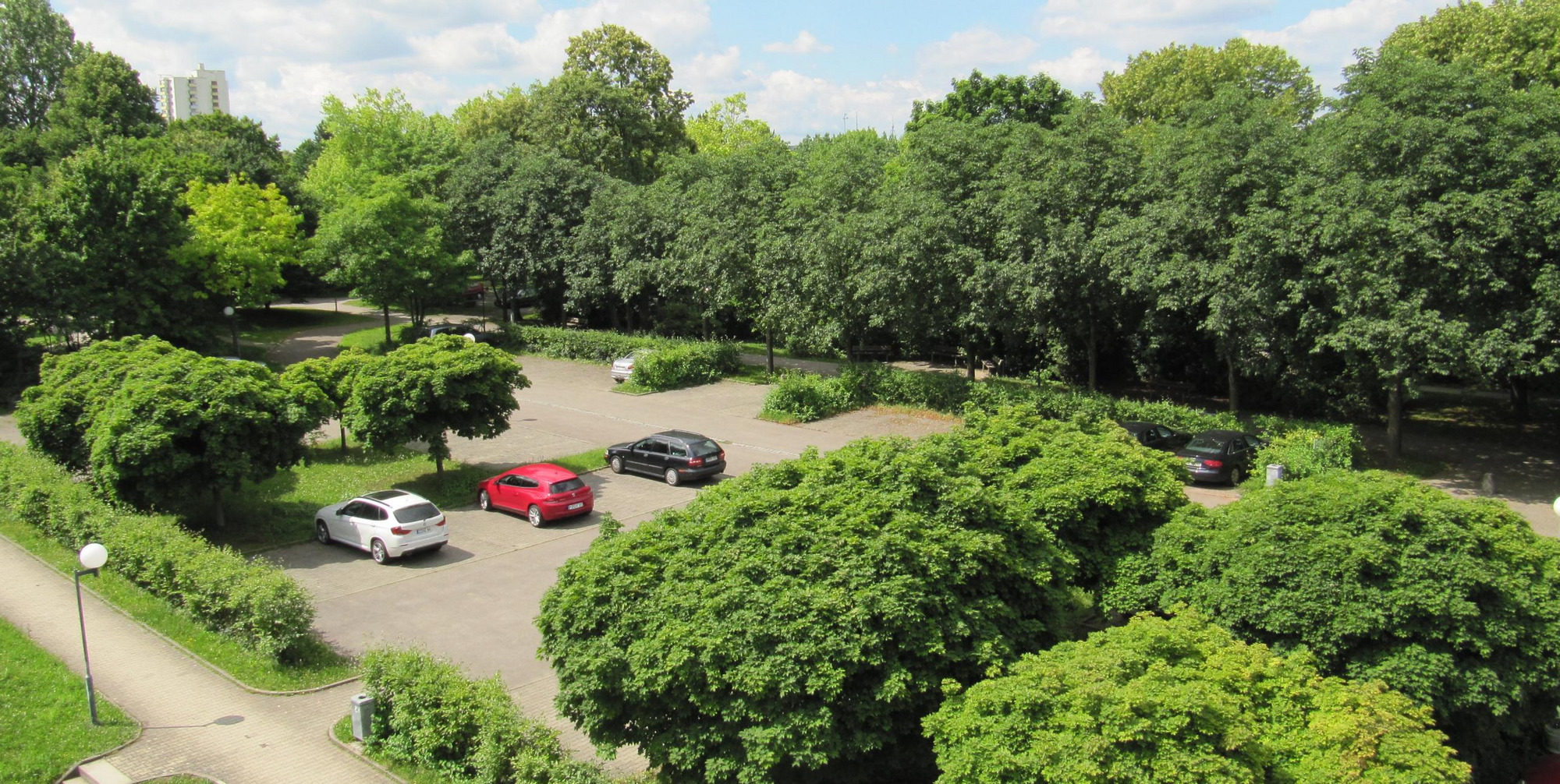 Parkplatz mit wenigen Autos, umgeben von grünen Bäumen und Büschen, teils bewölkter Himmel.