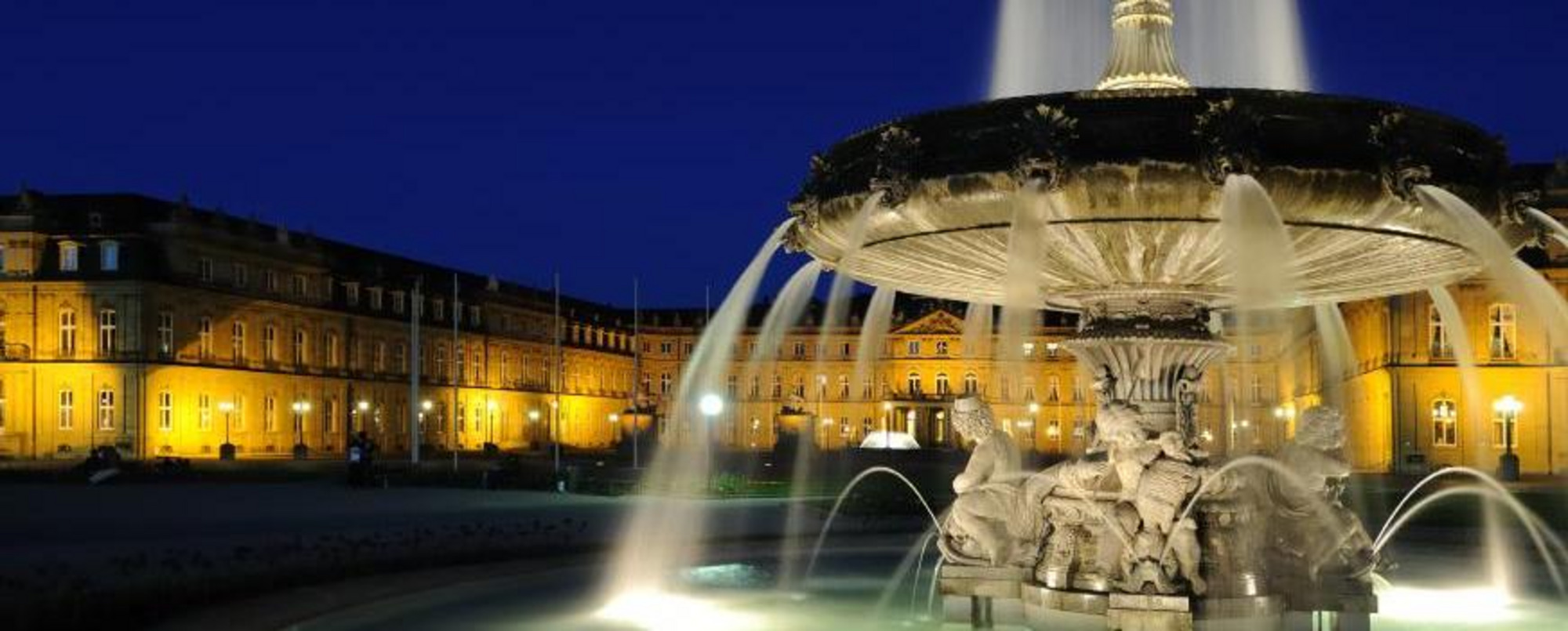 Nächtliche Ansicht eines beleuchteten Brunnens auf dem Schlossplatz Stuttgart mit Wasserstrahlen und historischer Kulisse im Hintergrund.