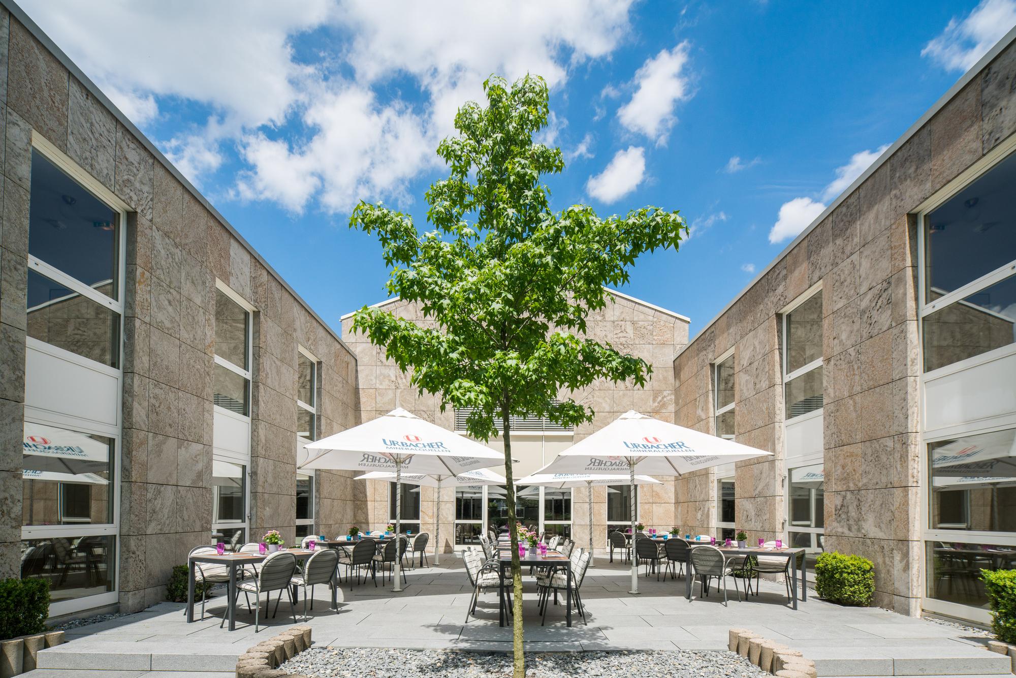 Terrasse mit großem Baum, Sitzgruppen unter weißen Schirmen, umgeben von Backsteingebäuden und blauem Himmel.
