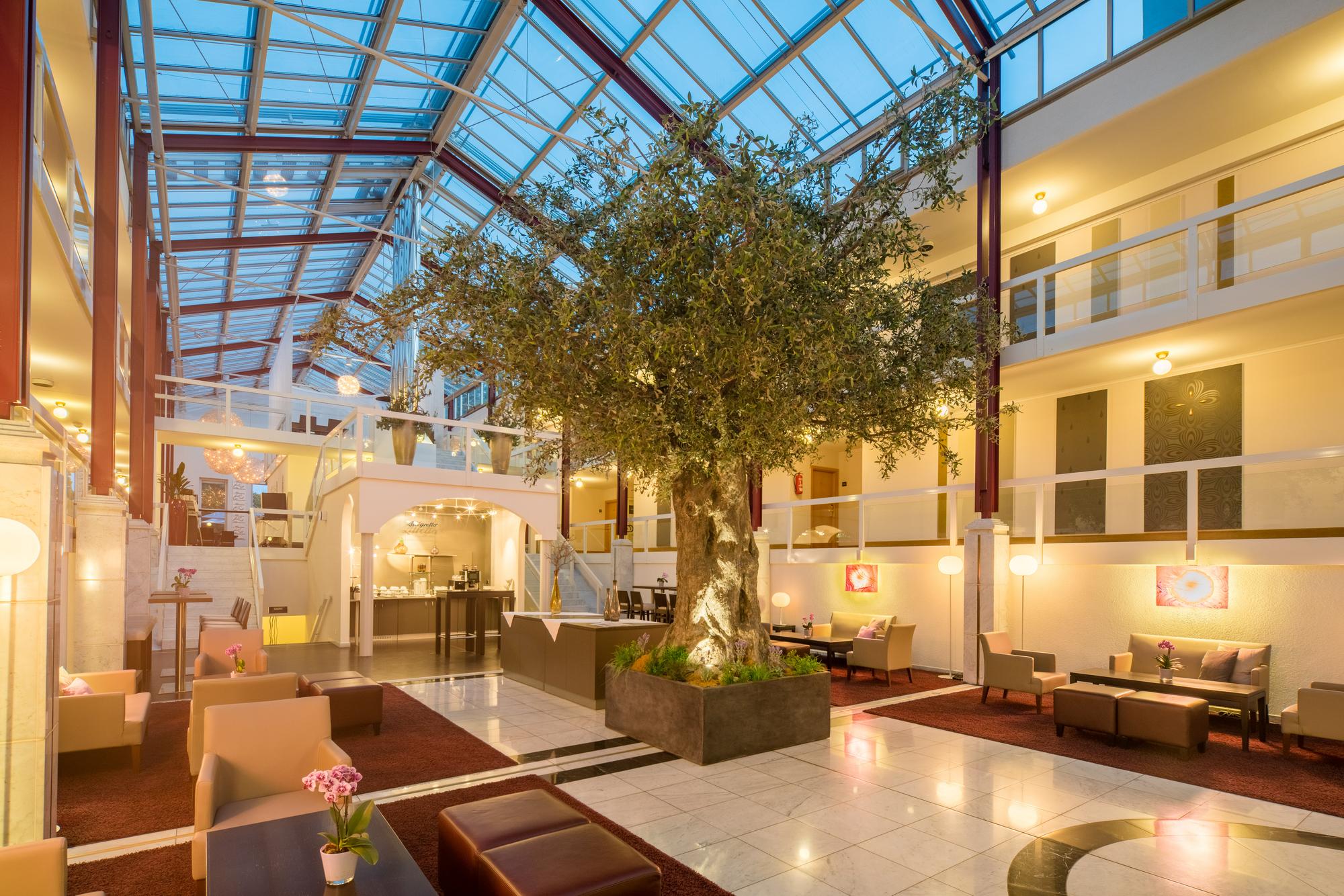 Großzügige Lobby mit Glasdecke, viel Tageslicht, Sitzgruppen, Pflanzen und einem zentralen Baum.