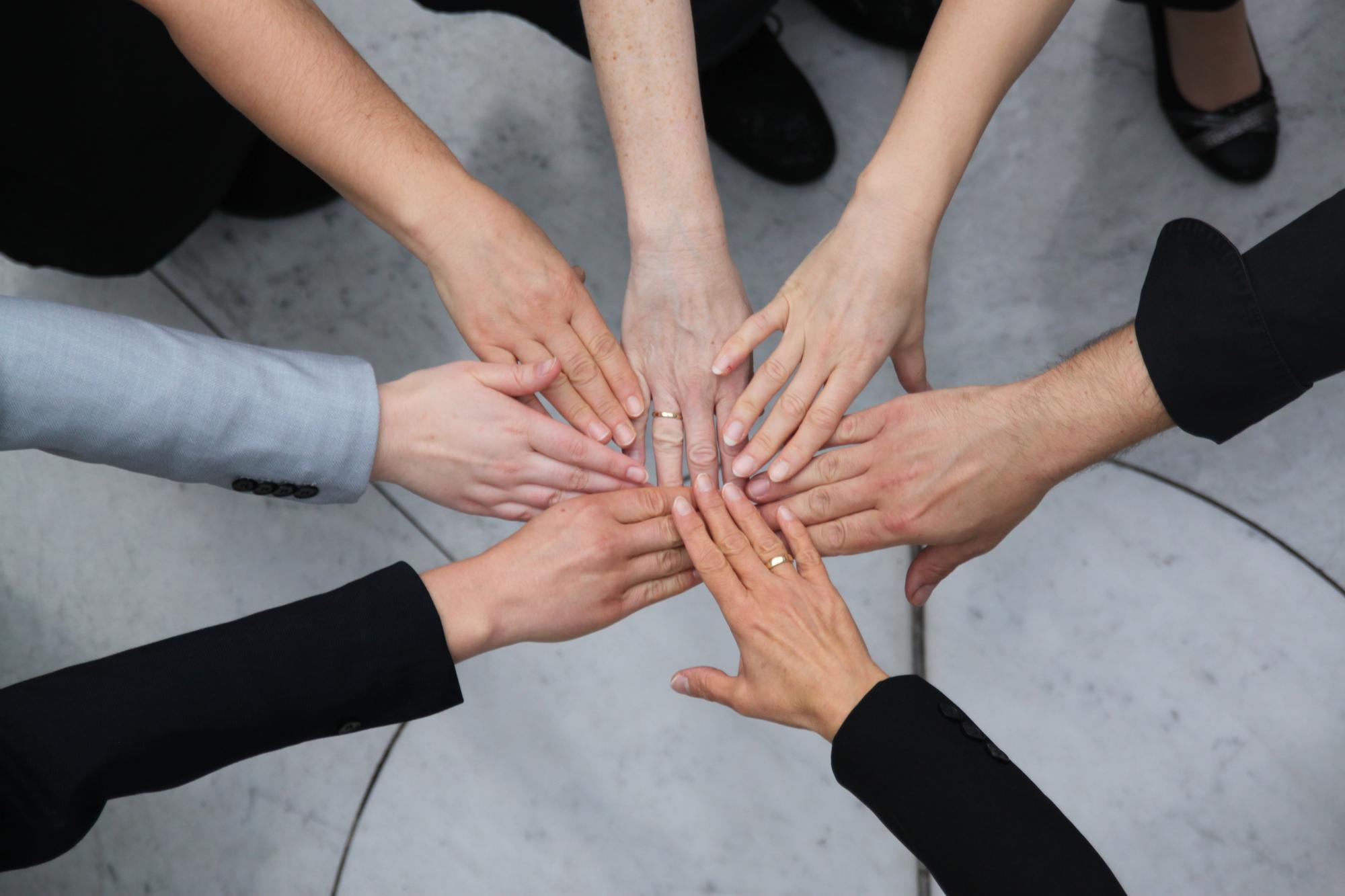 Gruppe von Personen legt die Hände übereinander in der Mitte zusammen – Symbol für Zusammenhalt und Teamwork.