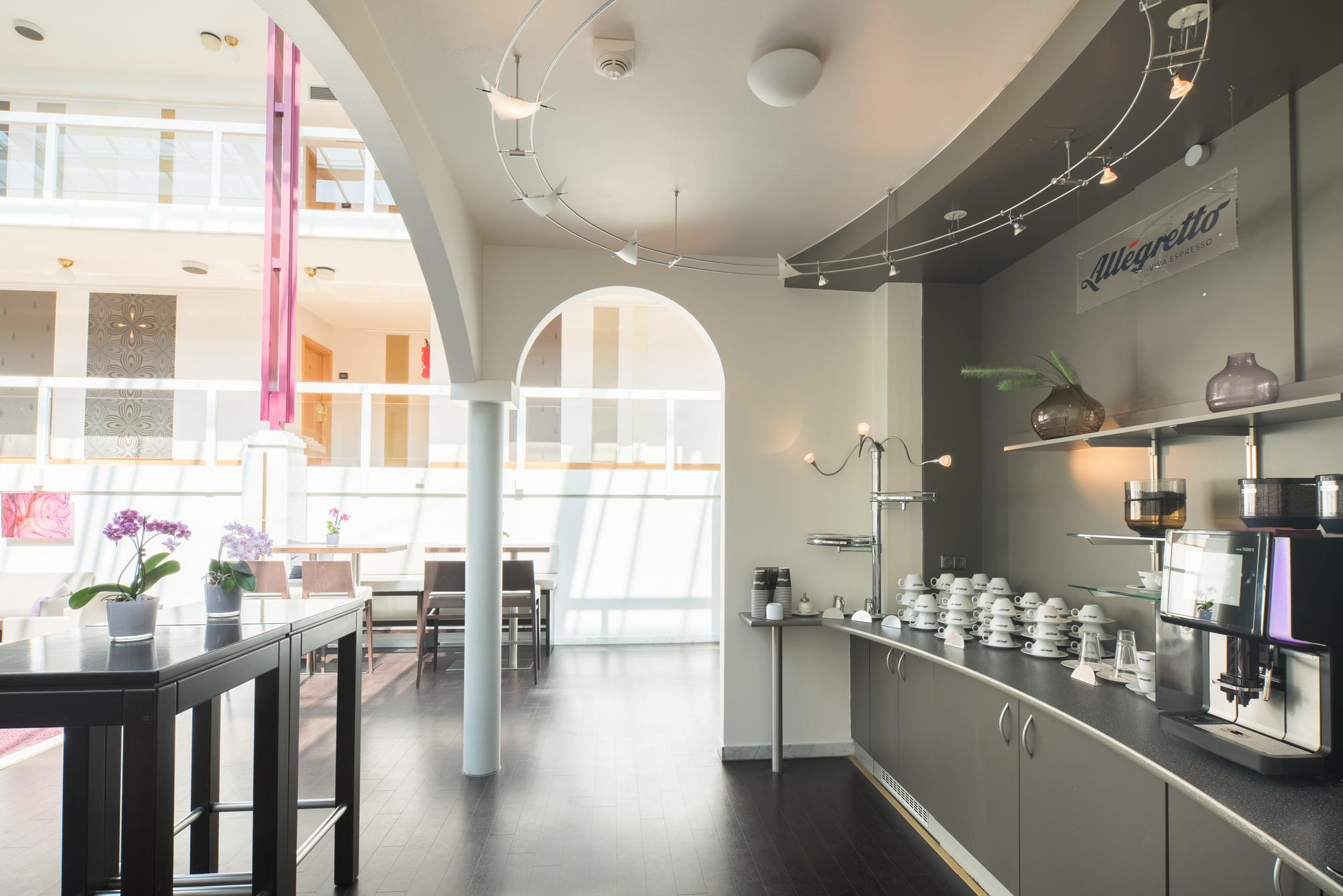 Moderne Kaffeebar mit Theke, Sitz- und Stehplätzen und großen Fenstern, durch die Tageslicht fällt.