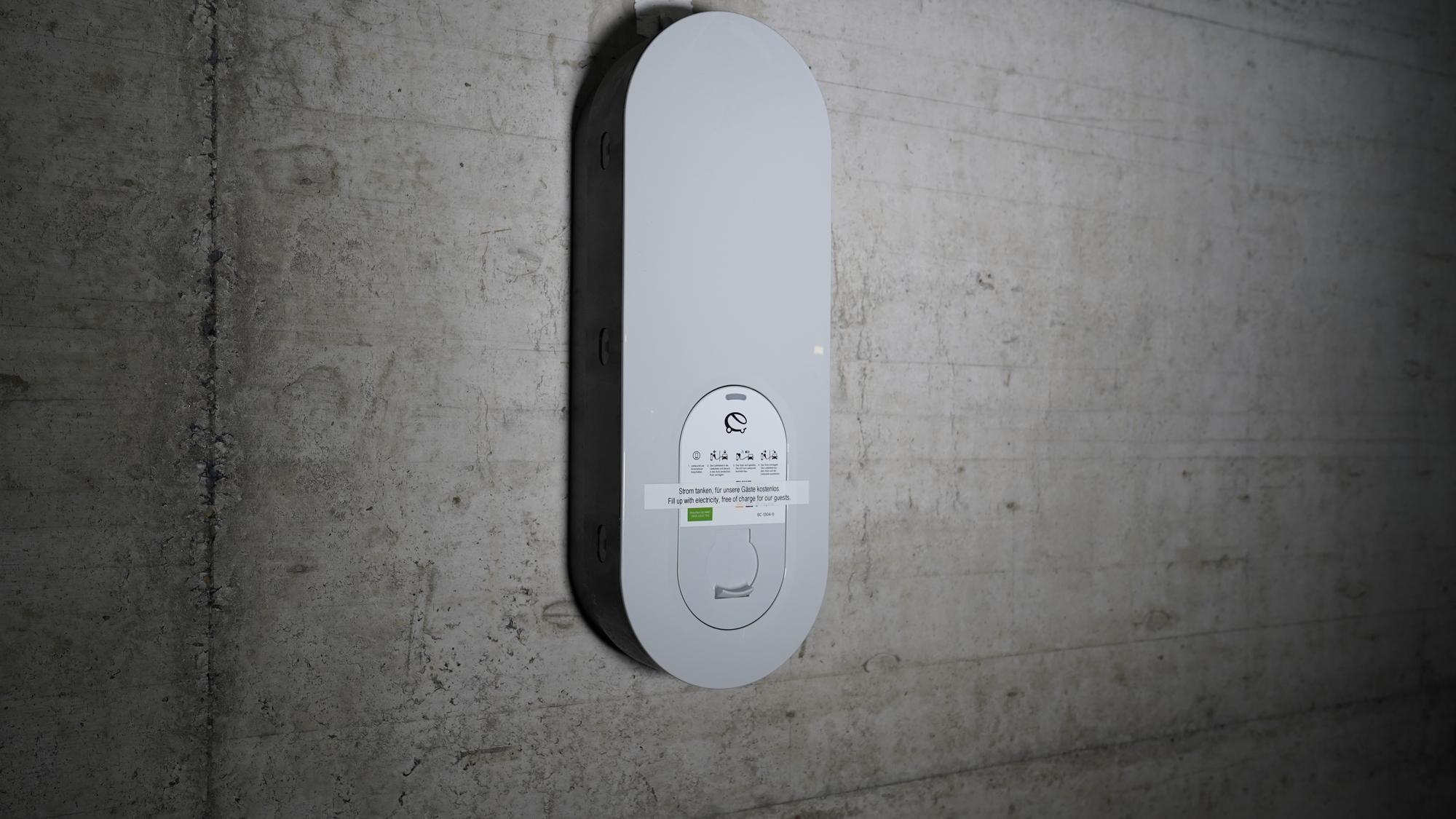 Nahaufnahme eines weißen, rechteckigen Objekts mit abgerundeten Ecken an grauer Wand – eine Ladestation für e-Autos.