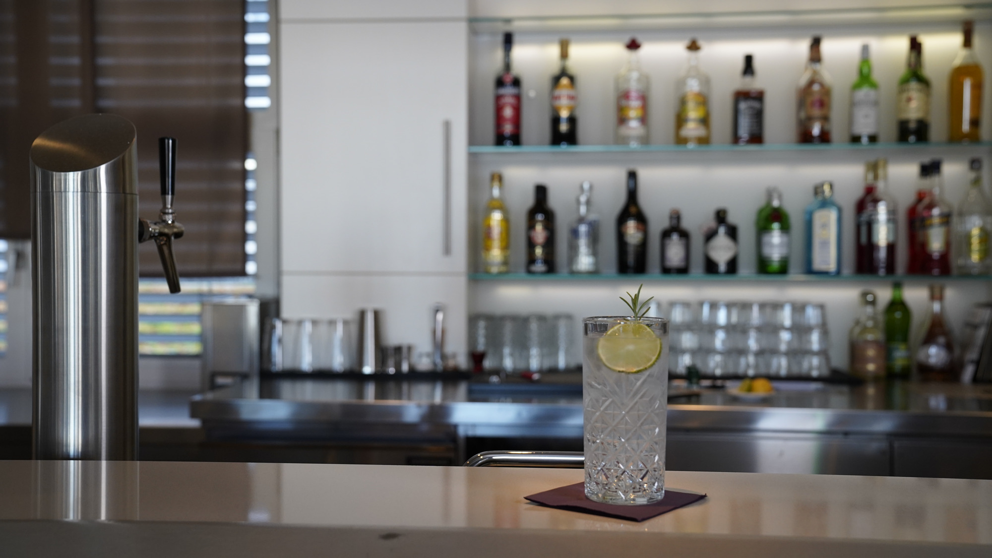 Bartresen mit Flaschenregal, Kühlschrank, Spüle, Gläsern und Gin Tonic auf roter Serviette.