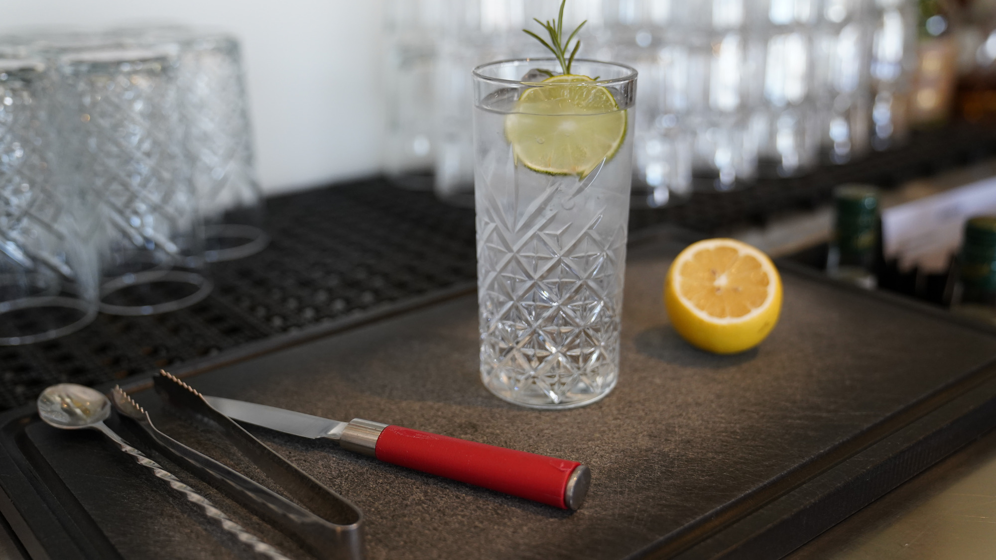 Küchenarbeitsplatte mit Besteck mit roten Griffen, Zitrone und Gin Tonic Glas und anderen Gläsern im Hintergrund.