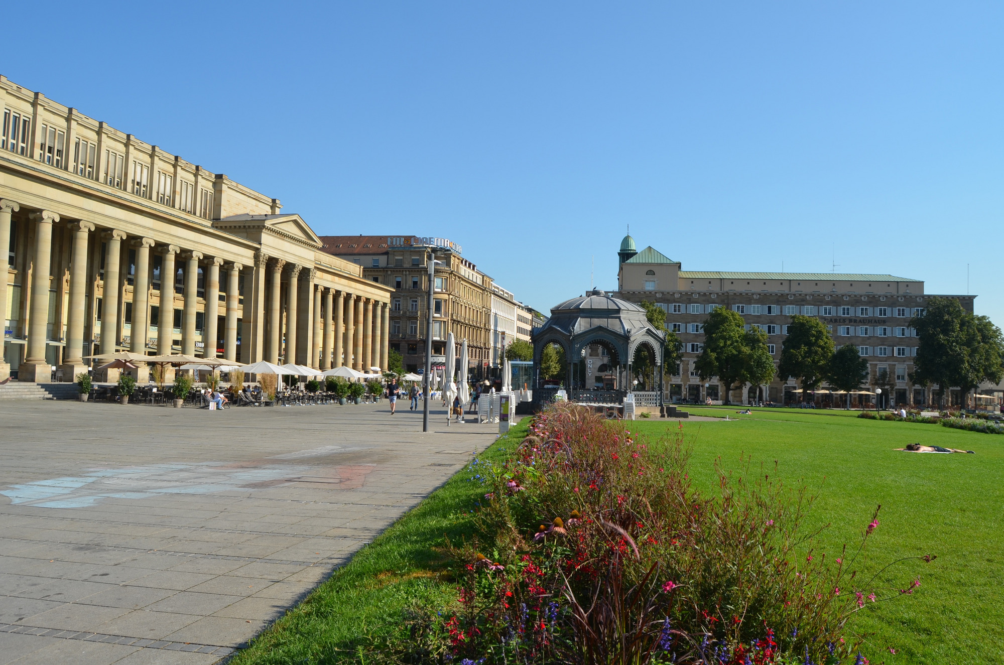 Königstraße Stuttgart mit historischen Gebäuden, Denkmal im Hintergrund, grüner Rasenfläche und blauem Himmel.