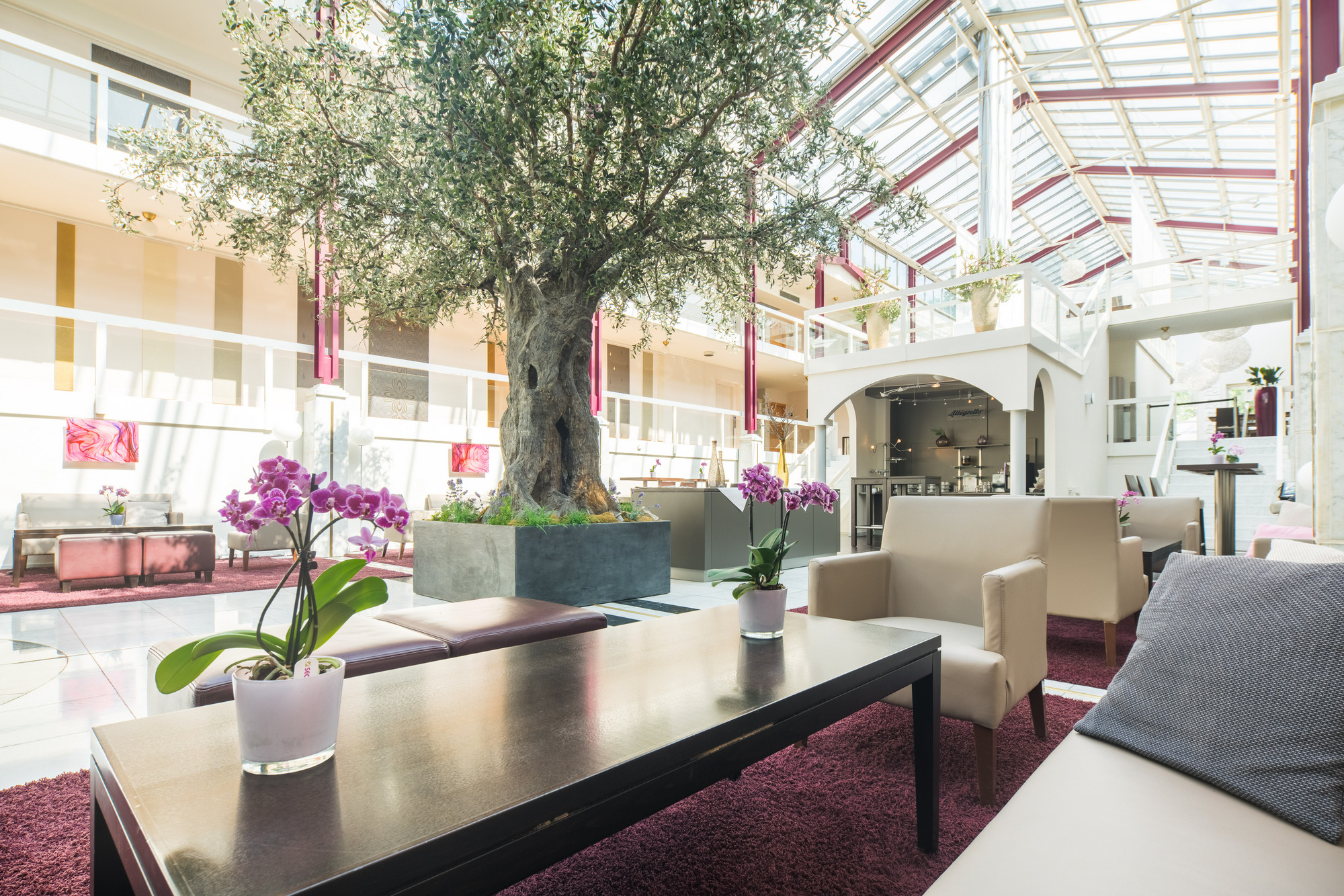Große, helle Lobby mit modernen Sofas, zentralem Baum, Pflanzen, hohen Decken und umlaufenden Balkonen.