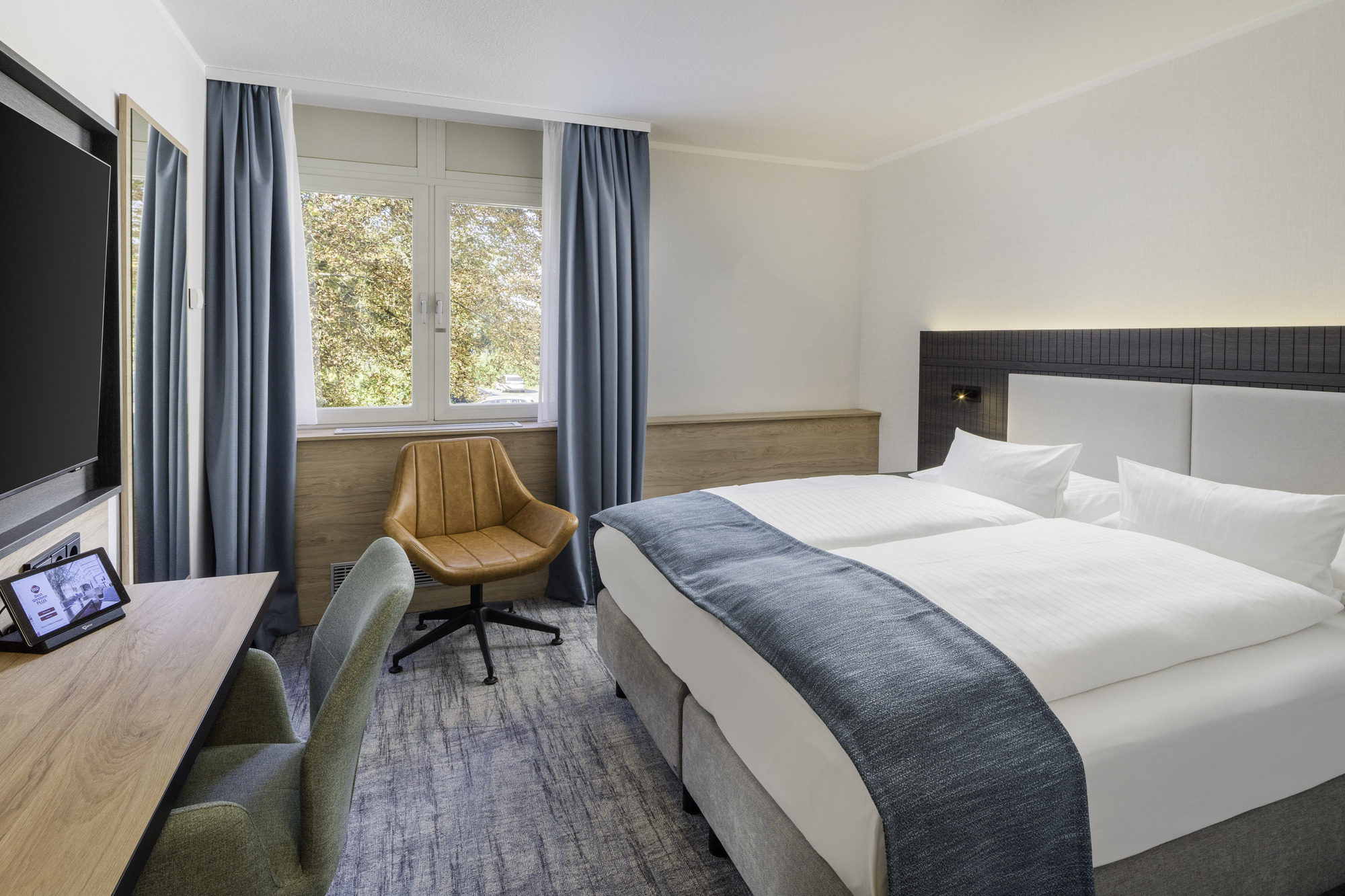 Modernes Hotelzimmer mit großem Bett, weißen Laken, türkisfarbenen Akzenten, orangefarbenem Sessel und Schreibtisch mit TV.