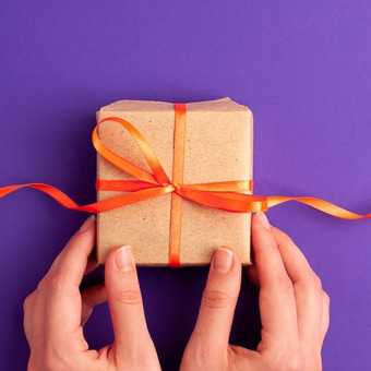 Zwei Hände halten Geschenk mit orangenem Band vor einem lila Hintergrund